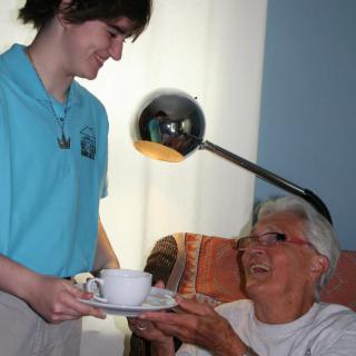 A caregiver assists an elderly person by serving tea in a home setting.