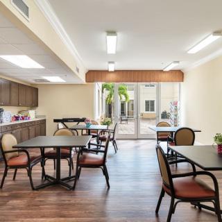 A well-lit dining area with multiple tables and chairs, featuring a kitchenette and large windows.