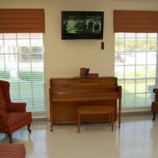 A cozy living room featuring a piano, two chairs, and a wall-mounted television between two large windows.