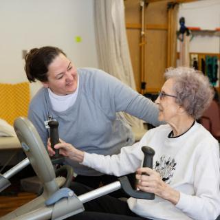 A caregiver assists an elderly woman with her exercise routine using fitness equipment.