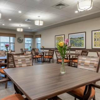 A cozy dining room with neatly arranged tables and chairs, decorated with flowers and artwork on the walls.