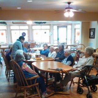 A group of seniors gathered around a table in a nursing home with a caregiver attending to them.
