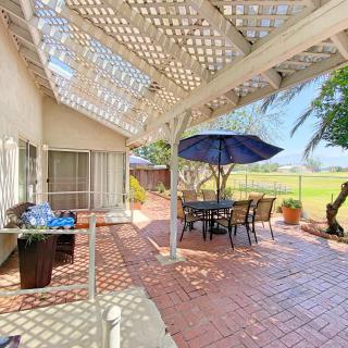 A sunny patio with a pergola, featuring a table and chairs under an umbrella, surrounded by plants and overlooking a fenced area.