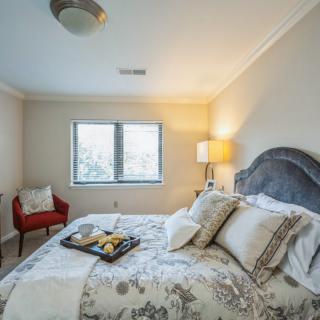 A cozy bedroom with a neatly made bed, decorative pillows, and a tray on the bed. A red chair and lamp are positioned by the window.