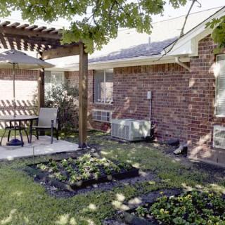 A cozy backyard with a pergola-covered patio, featuring outdoor seating and a small garden alongside a brick house.