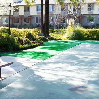 A sunny outdoor pathway near a residential area, surrounded by greenery and benches.