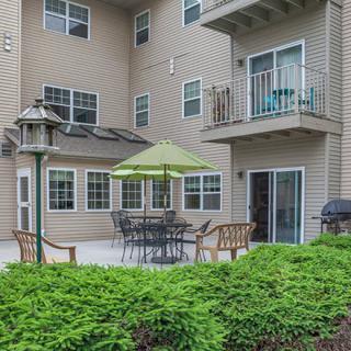 A cozy apartment patio with outdoor seating and a green umbrella, surrounded by lush greenery.