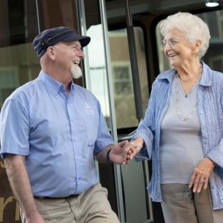 Two elderly individuals smiling and holding hands while standing outside.