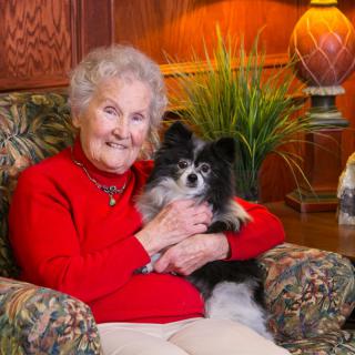 An elderly woman sitting on a chair, smiling and holding a small black and white dog in a cozy living room setting.