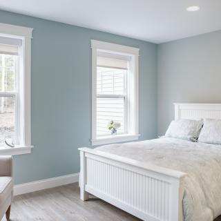 A minimalist bedroom featuring soft blue walls, a white bed frame, and ample natural light from two windows.