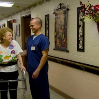 A healthcare worker assists an elderly woman using a walker in a nursing home hallway.