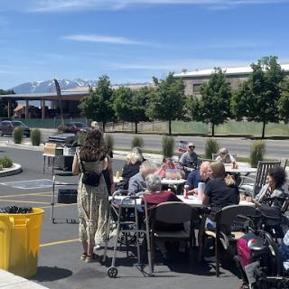 A community gathering outdoors on a sunny day, with people enjoying food at tables against a backdrop of mountains.