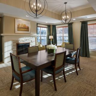 A modern dining room with a wooden table, upholstered chairs, and stylish chandeliers, featuring a fireplace and large windows with curtains.