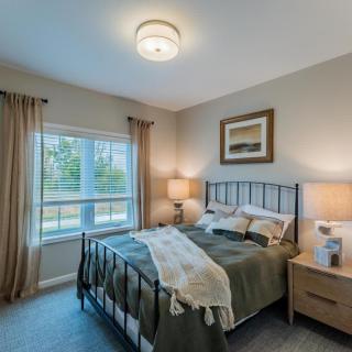 A cozy bedroom featuring a neatly made bed with green bedding, flanked by two nightstands and lamps, framed art on the wall, and natural light streaming in through a window with beige curtains.