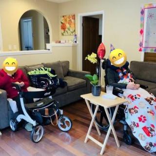 Two people smiling while sitting in wheelchairs in a cozy living room.