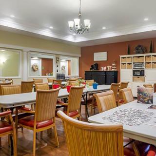 A well-lit community room with tables and chairs arranged for group activities, featuring a puzzle on one table and shelves containing various items.