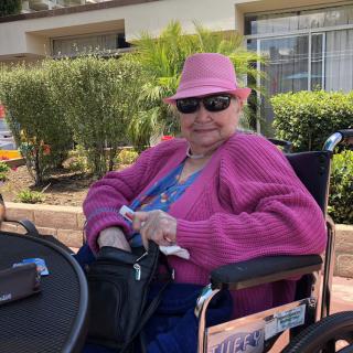 An elderly woman sitting in a wheelchair outdoors, wearing a pink hat and sunglasses.