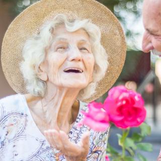 An elderly woman wearing a hat smiles joyfully in a garden, appreciating the bright pink flowers.
