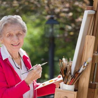 An elderly woman painting on a canvas outdoors, surrounded by art supplies and an easel.