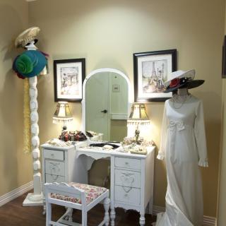 A vintage-themed room with a white vanity, a mannequin wearing a wedding dress, and various hats displayed.