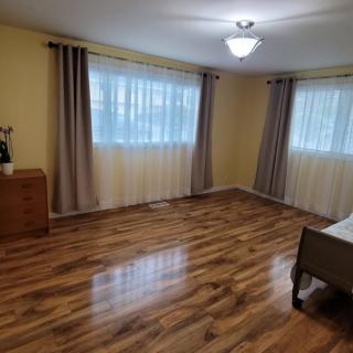A spacious bedroom with hardwood flooring, featuring large windows with curtains, a dresser, and a ceiling light fixture.