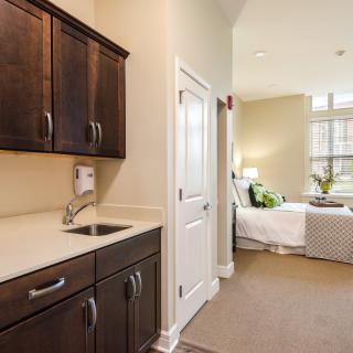 A modern apartment interior featuring a kitchenette with dark wooden cabinets leading to a cozy, well-lit bedroom.