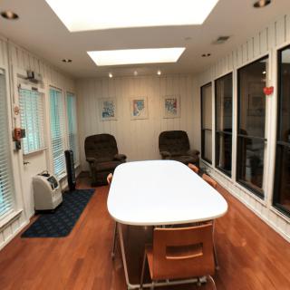 A well-lit room with a wooden floor, featuring a table and chairs, large windows, and skylights.