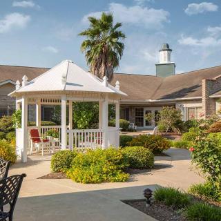 A serene garden featuring a white gazebo surrounded by lush greenery and a well-maintained pathway, set against the backdrop of a large building with a tower.