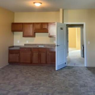 An empty room with a small kitchen area, featuring wooden cabinets and a carpeted floor.