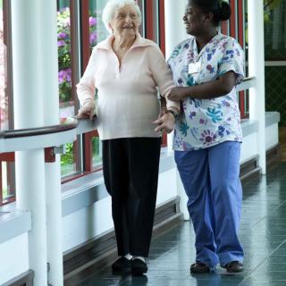 A nurse assists an elderly woman as they walk together in a corridor.