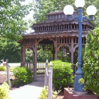 A wooden gazebo in a park surrounded by lush greenery and a lamp post.