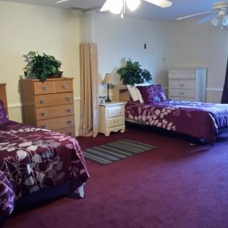 A cozy bedroom featuring twin beds with purple bedding, carpeted flooring, and wooden furniture.