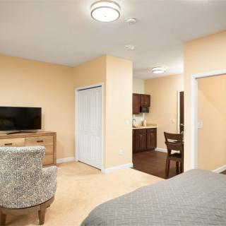 A cozy bedroom with a TV, dresser, and chair, adjacent to a kitchenette area.