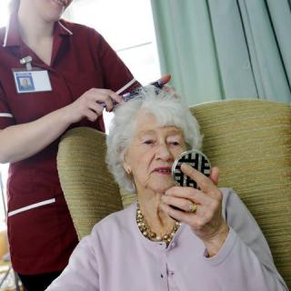 An elderly woman sitting in a chair uses a mirror as a caregiver brushes her hair.