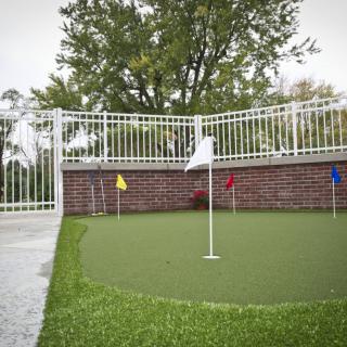 A small putting green with multiple colored flags, surrounded by a white fence in an outdoor setting.
