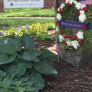 A memorial wreath is displayed in a garden setting, surrounded by greenery and flowers.