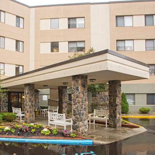 The image shows the entrance of a multi-story building with stone columns and benches, surrounded by landscaped areas.