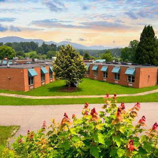 A scenic view of a school building surrounded by lush greenery and mountains under a cloudy sky.