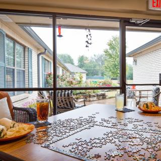 A cozy indoor setting with a puzzle on the table, along with food and drinks, overlooking a courtyard through large windows.
