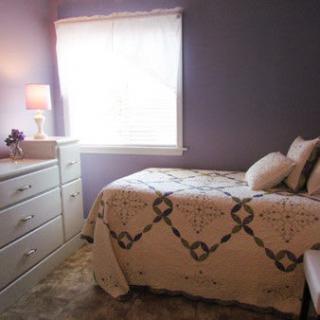 A cozy bedroom with purple walls, a neatly made bed, and a dresser beside a window letting in natural light.