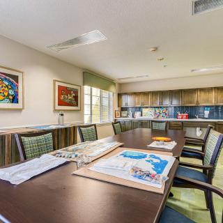 A spacious conference room with a long table, chairs, vibrant artwork on the walls, and a kitchenette in the background.
