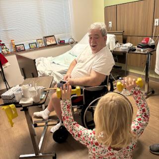 A child helps an elderly patient in a wheelchair with some light exercise while in a hospital room.