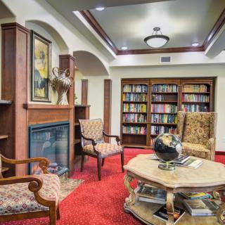 A cozy library room featuring bookshelves, comfortable chairs, a table with a globe, and a fireplace under warm lighting.