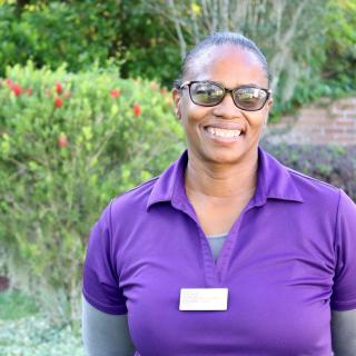 A person wearing a purple shirt and glasses, smiling in an outdoor setting.