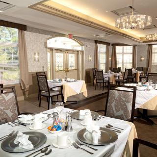 An elegant dining room with neatly set tables and a chandelier, featuring large windows and soft lighting.