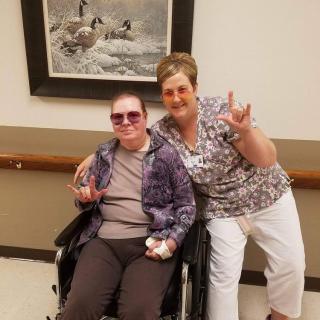 Two people indoors, one in a wheelchair, both smiling and making hand gestures in front of a framed artwork.