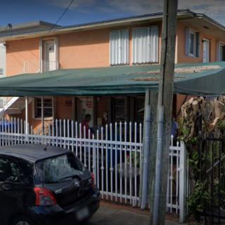 A two-story residential house with a car parked in front and a white fence surrounding the property.