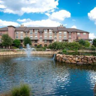 A scenic view of a lakeside hotel with a fountain and lush greenery under a blue sky.