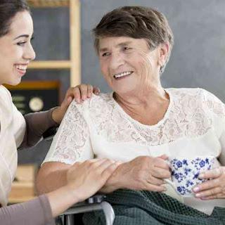 A caregiver and an elderly woman share a joyful moment over coffee in a warm, indoor environment.