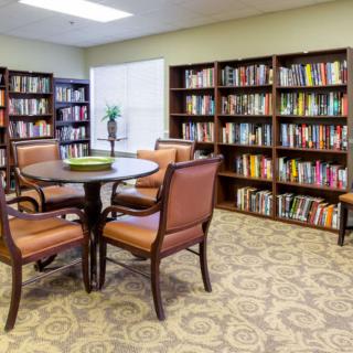 A cozy library room with bookshelves filled with books, a round table, and comfortable chairs on a patterned carpet.